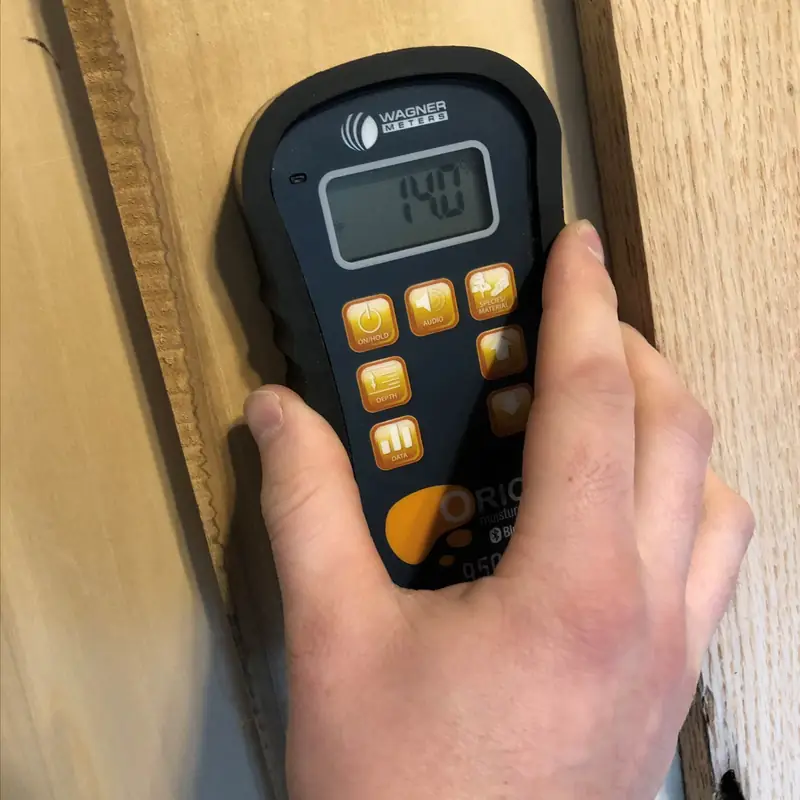 Käsi painaa puun kosteusmittarin laudan pintaan; näytössä lukema 14,0.